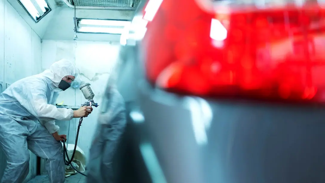 Mann in weißem Schutzanzug besprüht ein Auto in Lackierkabine mit Lack