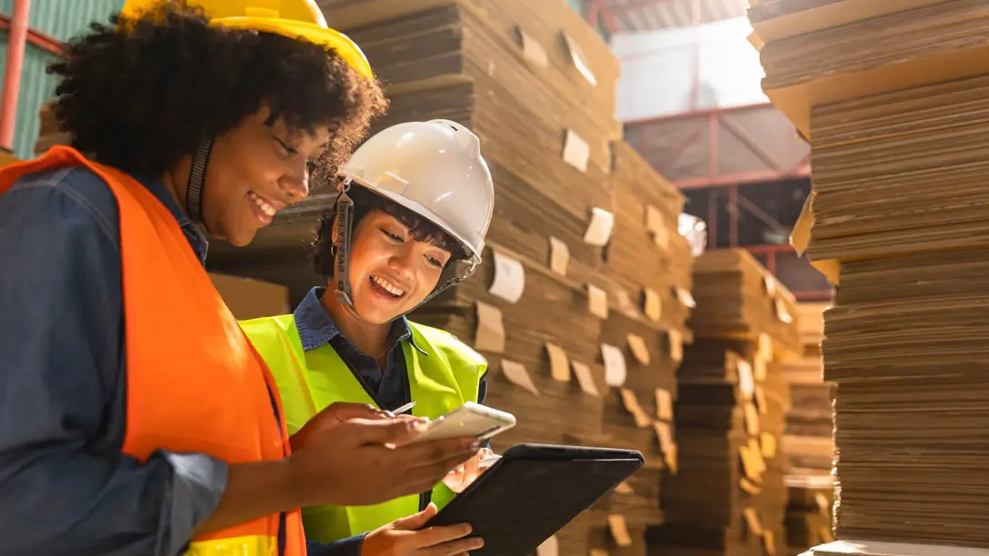 Arbeiterinnen mit Helmen schauen auf Tablet in Lagerhalle